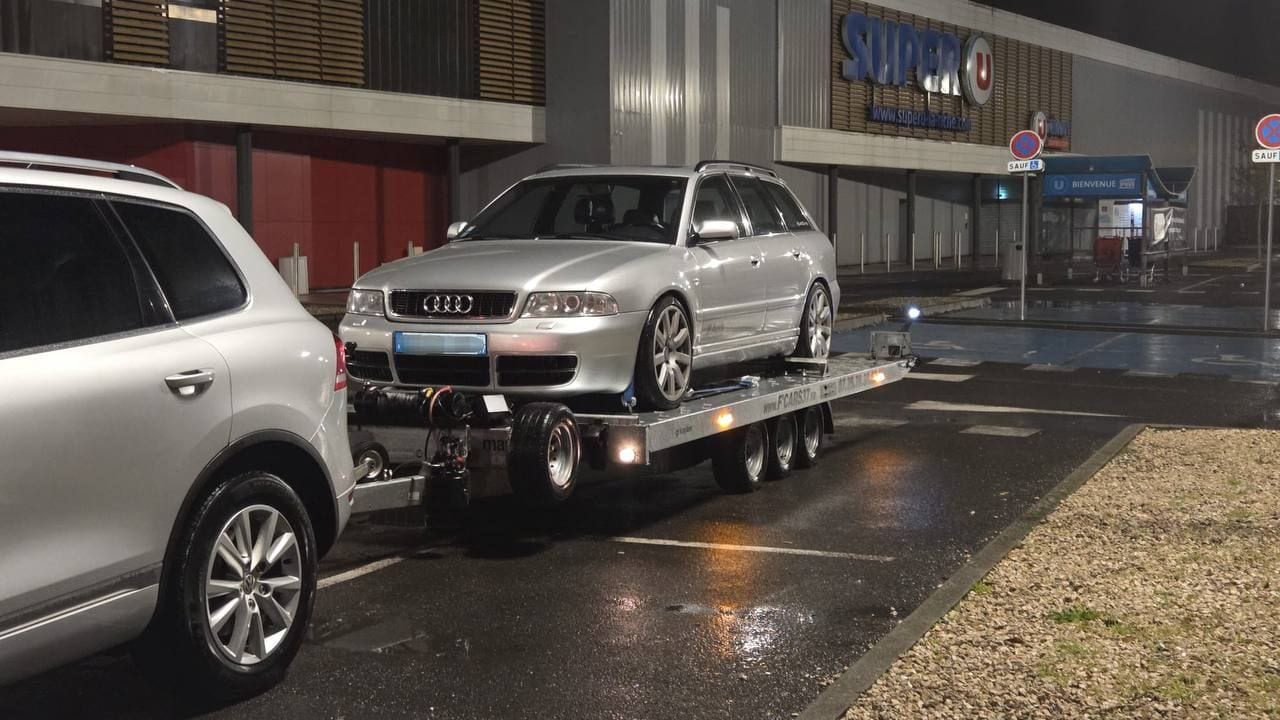 Break Audi chargé de nuit sous la pluie sur une remorque plateau, sur un parking de supermarché