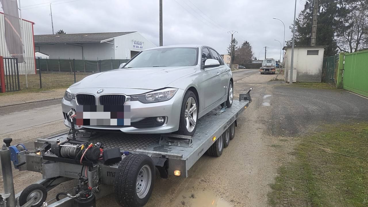 BMW Série 3 accidentée à l'avant, treuillée sur une remorque plateau devant un garage