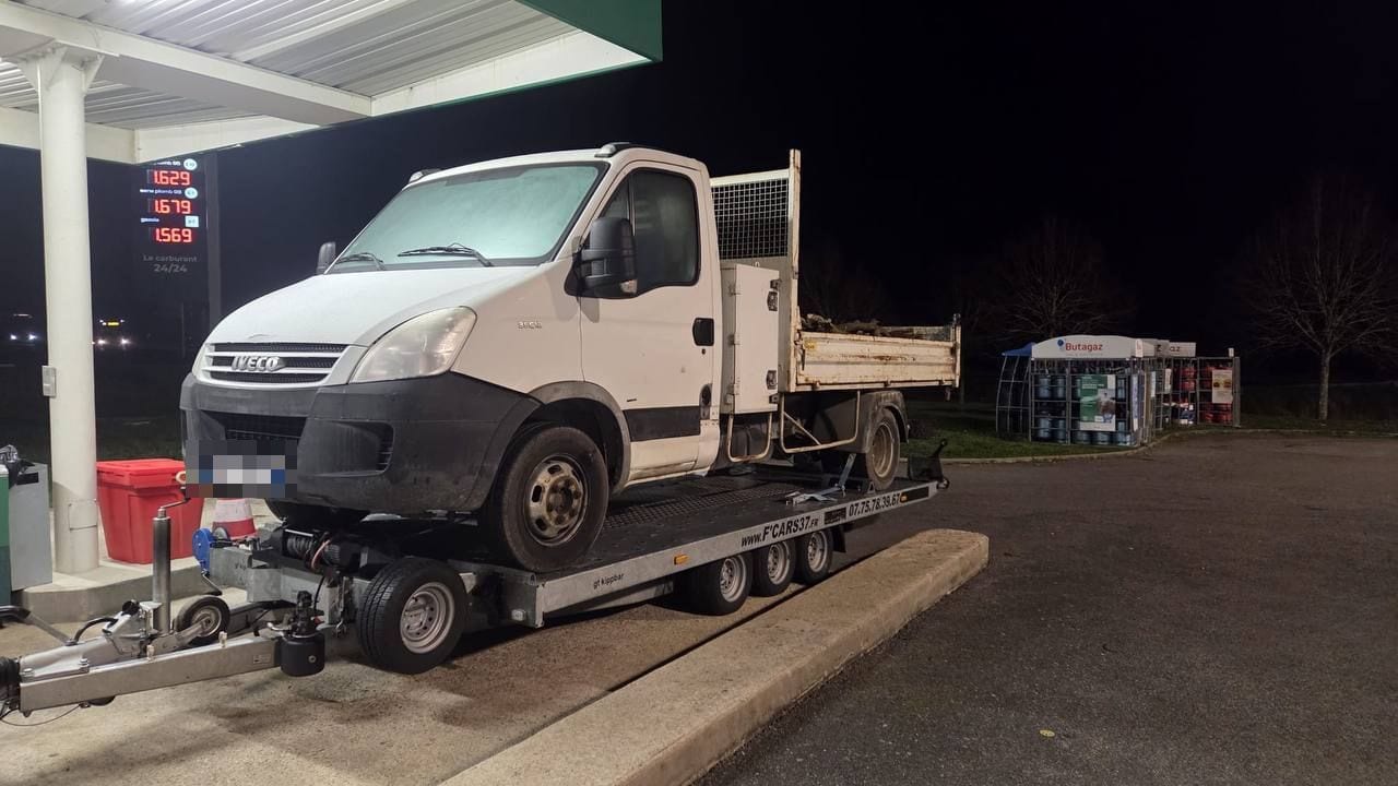 Camion benne Iveco Daily arrimé sur une remorque plateau, de nuit dans une station-service