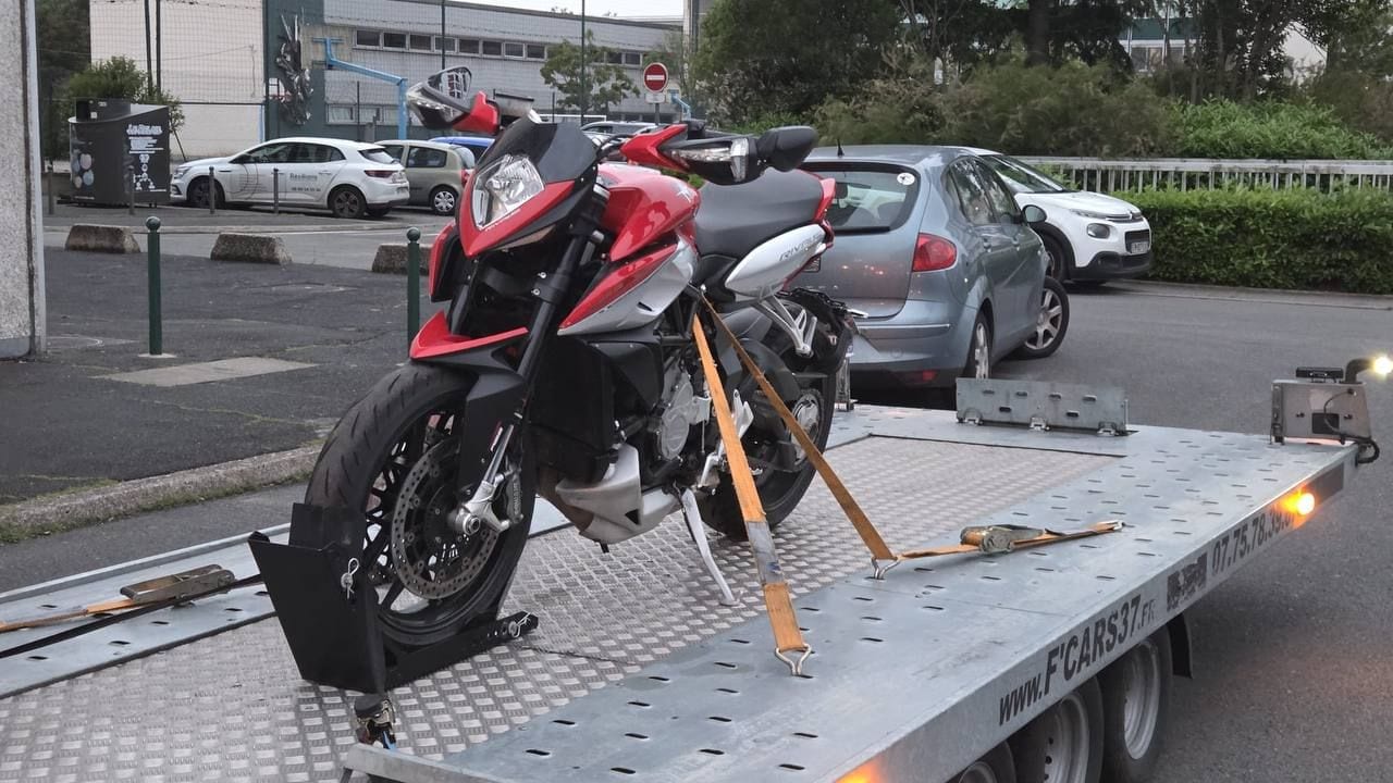Moto MV Agusta Rivale rouge maintenue debout sur une remorque plateau par un bloque-roue et des sangles croisées